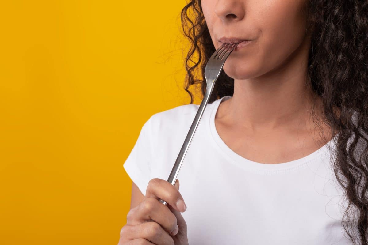 Tasty Meal. Close up of hungry young lady holding fork in her mouth, biting and dreaming about delicious healthy food, choosing something to eat isolated on yellow orange studio wall, free copy space