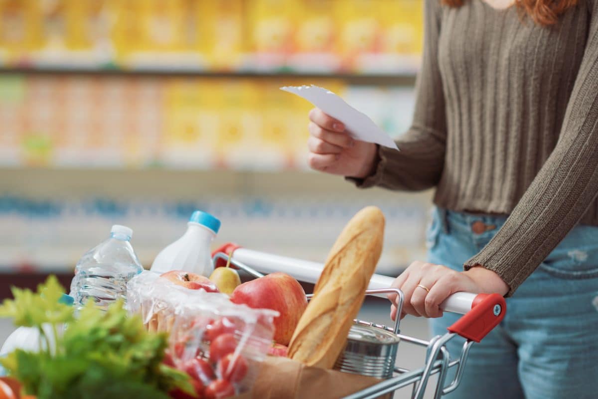 Young woman doing grocery shopping at the supermarket, she is pushing a full trolley and holding a shopping list, close up